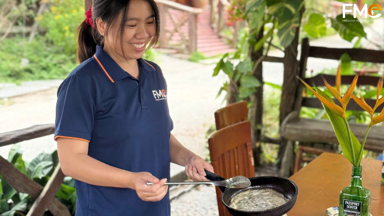 A smiling FME Travel staff member interacting with the cooking setup, showcasing the friendly and authentic "Rustic Hospitality" of the Mekong.