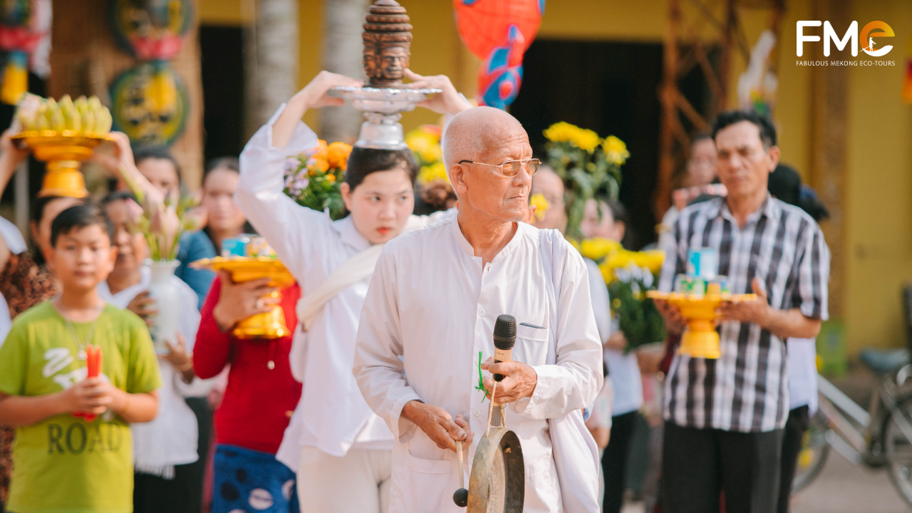 An elderly Khmer man in a white shirt and glasses striking a gong to lead a procession, followed by people carrying offerings and flowers.