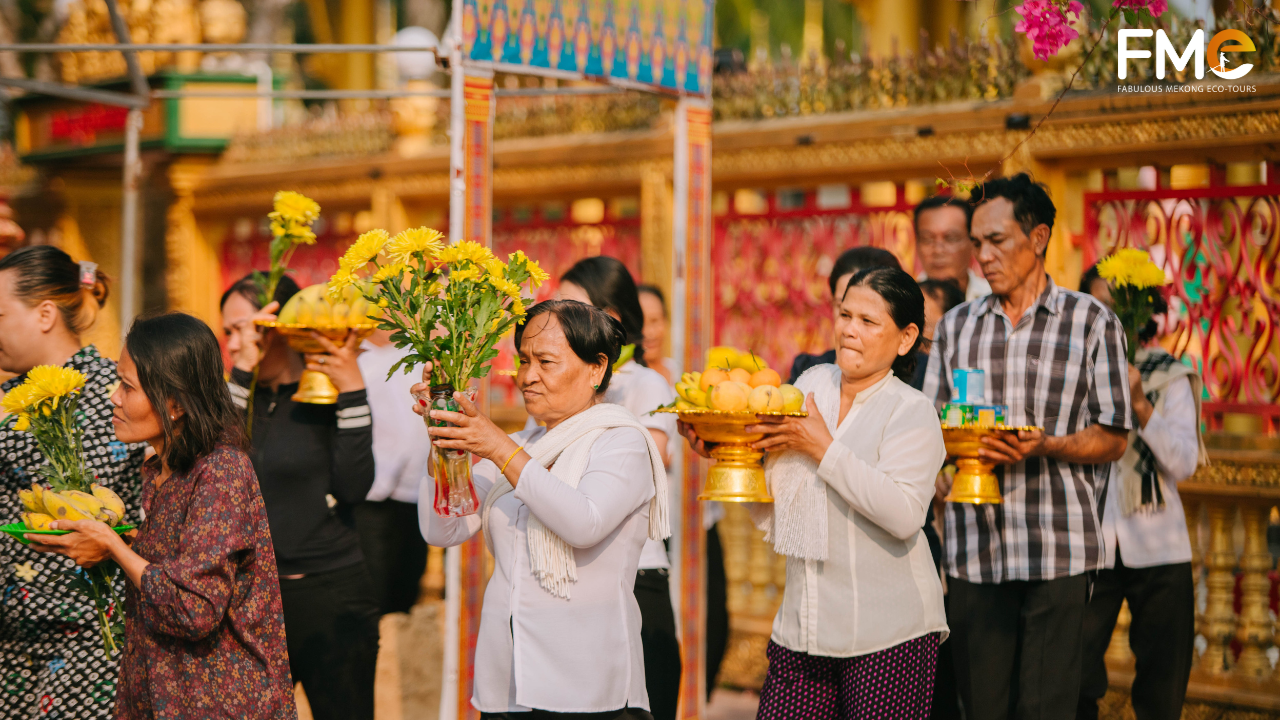 An elderly woman in white carrying a vase of bright yellow chrysanthemums and a tray of fruit on her head during an offering procession at a Khmer pagoda.