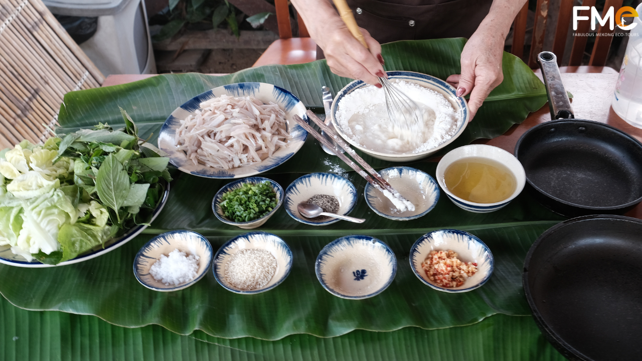 Traditional ingredients for making Lonton batter cake, including flour, fresh "long tong" fish, garden herbs, and spices arranged on a green banana leaf in a Mekong Delta cooking class.