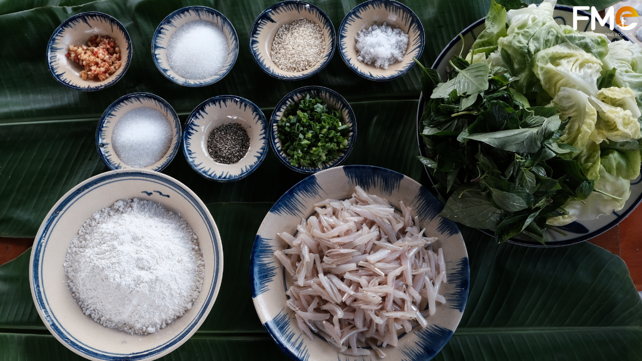 A top-down view of traditional cooking ingredients including flour, fresh herbs, spices, and "lòng tong" fish arranged on a green banana leaf.