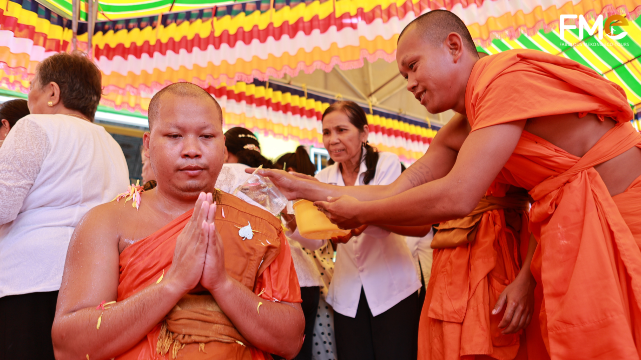 A monk sitting in prayer while being bathed with floral-scented water by another monk and local people, a ritual of purification and blessings.
